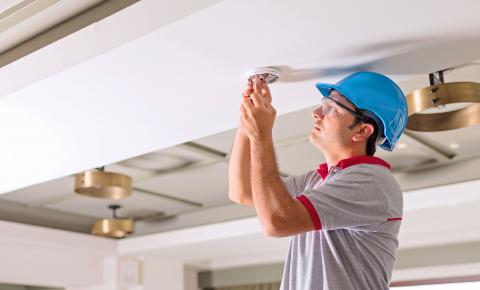 Fire alarm technician installs wired detectors in a ceiling.